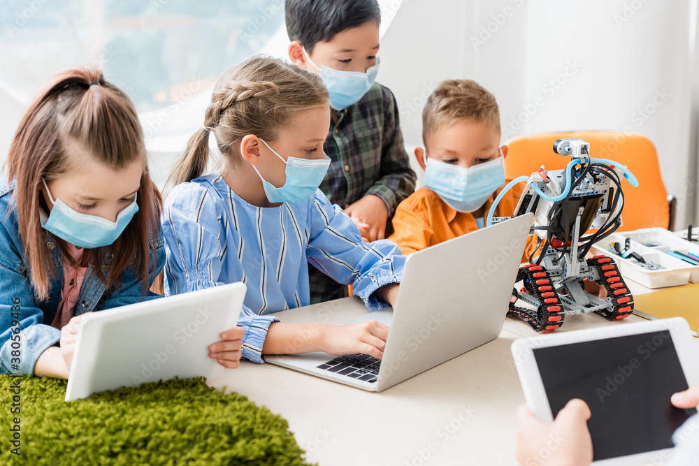 Selective focus of multiethnic children in medical masks using digital tablet and laptop near teacher and robot in school Selective focus of multiethnic children in medical masks using digital tablet and laptop near teacher and robot in school