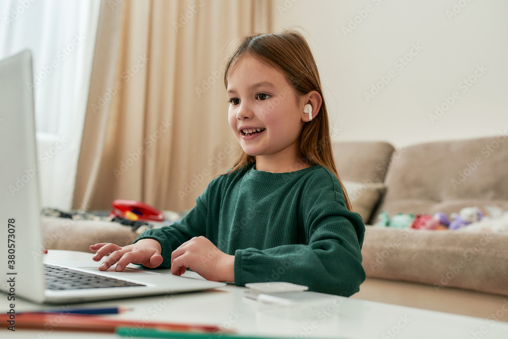 A cute small girl enjoying doing her distance education hometask and smiling while sitting alone in front of a laptop at the table A cute small girl enjoying doing her distance education hometask and smiling while sitting alone in front of a laptop at the table
