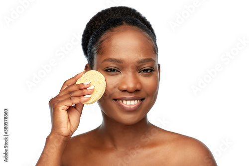 beauty, people and skincare concept - happy smiling young african american woman with bare shoulders cleaning face with exfoliating sponge over white background