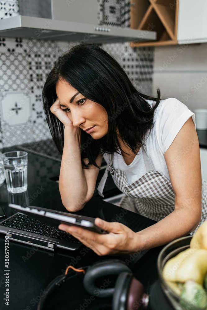 Unhappy and worried middle age woman sitting alone in her apartment and using laptop computer or tablet. She is bored because social distancing during pandemic quarantine. Unhappy and worried middle age woman sitting alone in her apartment and using laptop computer or tablet. She is bored because social distancing during pandemic quarantine.