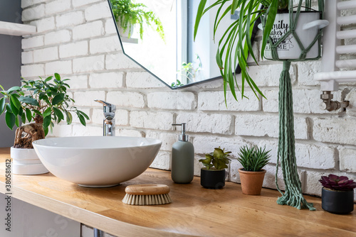 Bathroom with white bricks, green plants, stylish mirror, ceramic wash basin on wooden countertop. Industrial interior in apartment at home.