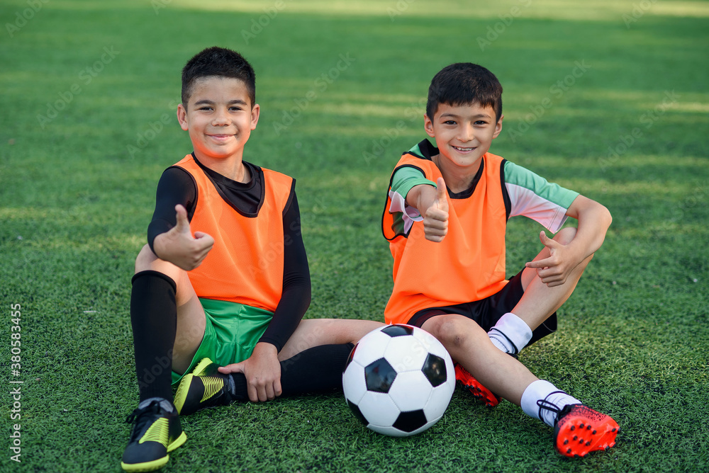 Joyful teenage male soccer players show ok sign resting on artificial turf of football field after intensive training. Joyful teenage male soccer players show ok sign resting on artificial turf of football field after intensive training.
