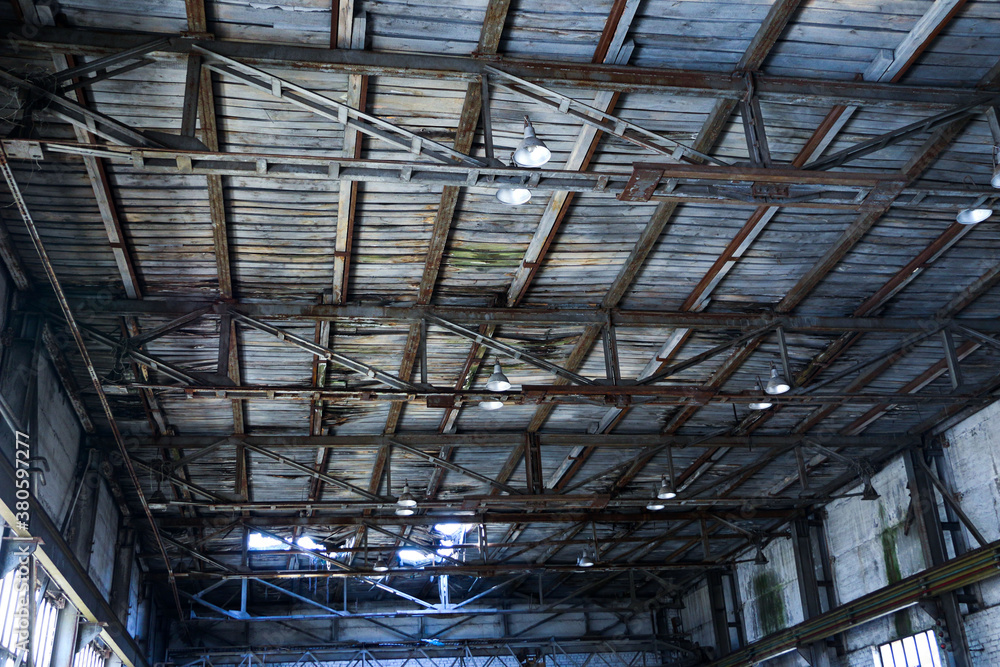 An old, decayed wooden ceiling of an abandoned factory, with large iron lamps, rusted steel structures, cobwebs and mildew.
