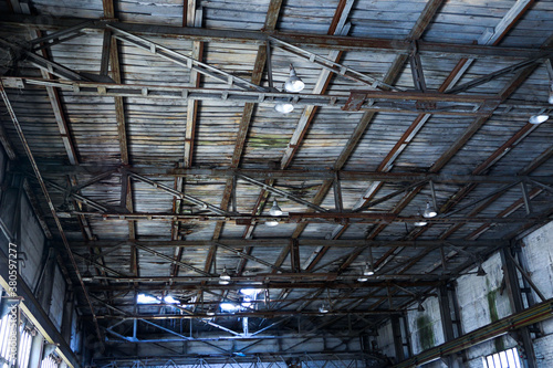 An old, decayed wooden ceiling of an abandoned factory, with large iron lamps, rusted steel structures, cobwebs and mildew.
