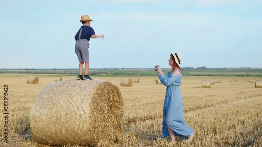 family holiday, cheerful female parent takes photograph on retro camera of her adorable male child on haystack in field after harvesting against sky family holiday, cheerful female parent takes photograph on retro camera of her adorable male child on haystack in field after harvesting against sky