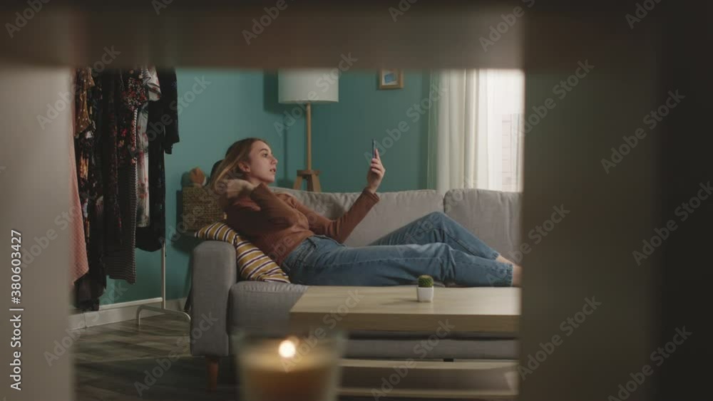 The young lady in blue jeans and a brown sweater lying on the sofa and talking via video phone communication. Directions camera right. In the foreground a mezzanine with a lighted candle. The young lady in blue jeans and a brown sweater lying on the sofa and talking via video phone communication. Directions camera right. In the foreground a mezzanine with a lighted candle.