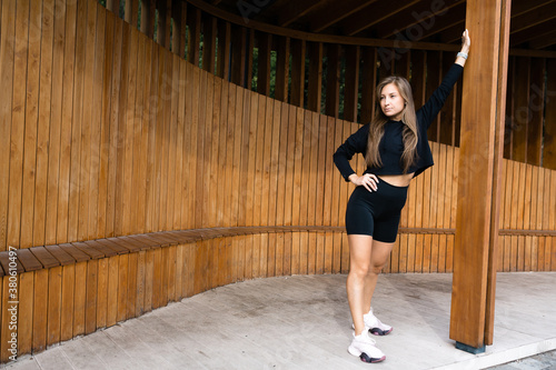 Girl in black sportswear stands and leans on a wooden support in the park