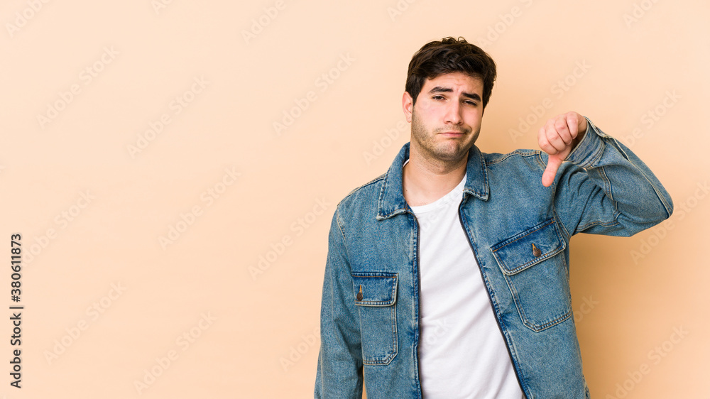 Young man isolated on beige background showing a dislike gesture, thumbs down. Disagreement concept. Young man isolated on beige background showing a dislike gesture, thumbs down. Disagreement concept.