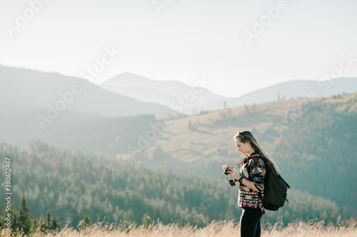 Hipster young woman with a backpack enjoying sunset on foggy mountain. Tourist girl hiker looking sunlight on trip in country. Female traveler photographing mountain at sunrise. Mockup text. © Serhii