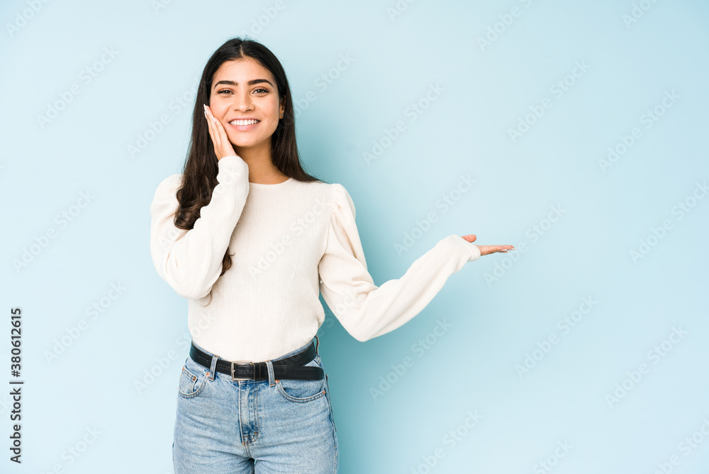 Young indian woman isolated on blue background holds copy space on a palm, keep hand over cheek. Amazed and delighted. Young indian woman isolated on blue background holds copy space on a palm, keep hand over cheek. Amazed and delighted.