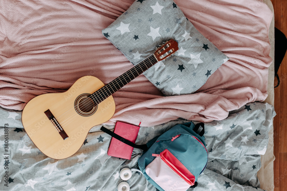 Student's room after study. Guitar, backpack, heaphones and notebook on the bed.
