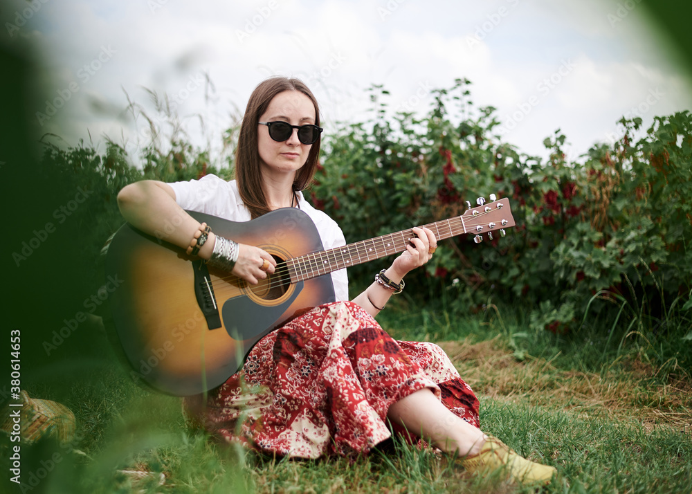 Young brunette hippie woman, wearing boho style clothes, sitting on green grass, holding guitar. Indie musician relaxing on green currant field on sunny summer day. Eco tourism concept.