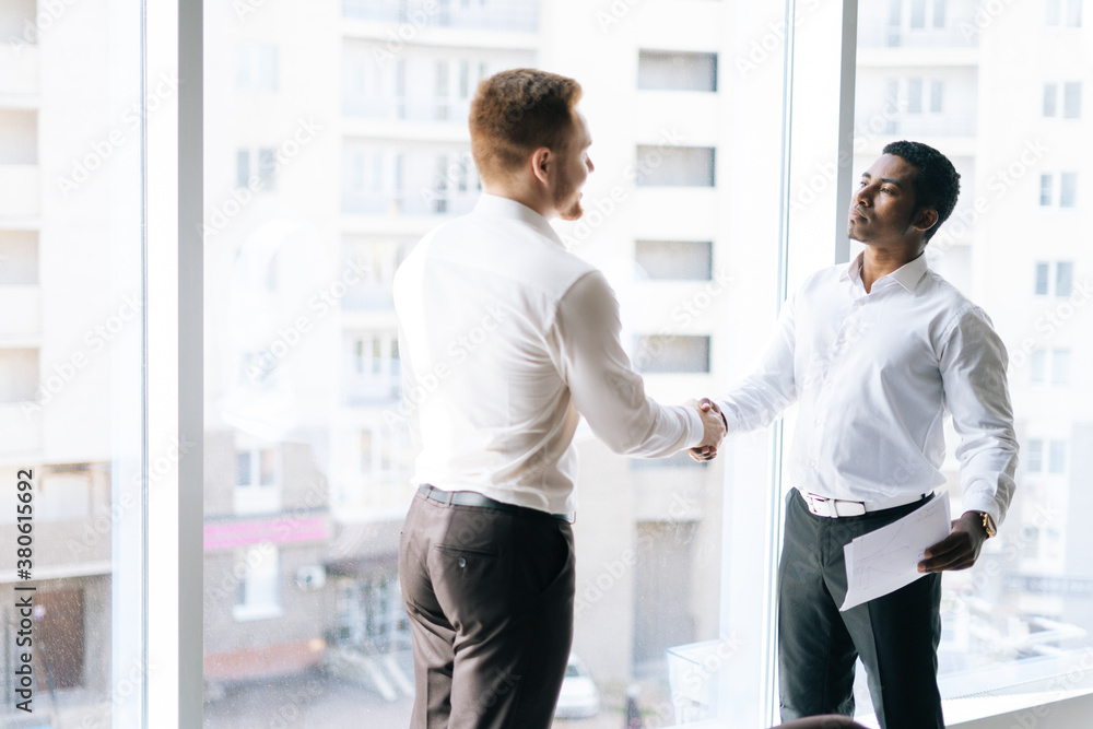 Caucasian businessman and serious African American businessman agreed contract and shaking hands in conference room background of window. Concept of interracial cooperation. Caucasian businessman and serious African American businessman agreed contract and shaking hands in conference room background of window. Concept of interracial cooperation.
