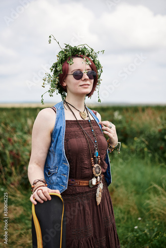 Young hippie woman with short red hair, wearing boho style clothes, sunglasses and flower wreath, standing on green field, holding guitar. Indie musician in countryside. Eco tourism concept.