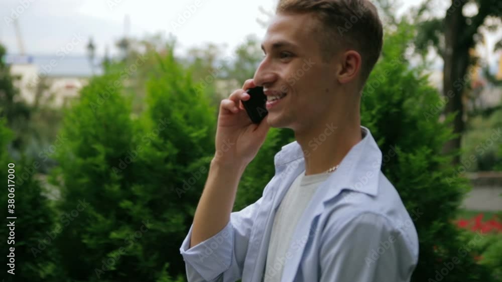 handsome blond guy walks along green city park and talks on modern black smartphone with smile slow motion closeup