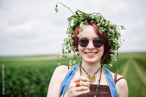 Young hippie woman with short red hair, wearing boho style clothes and flower wreath, standing on green currant field, posing for picture. Close-up female portrait on natural background.