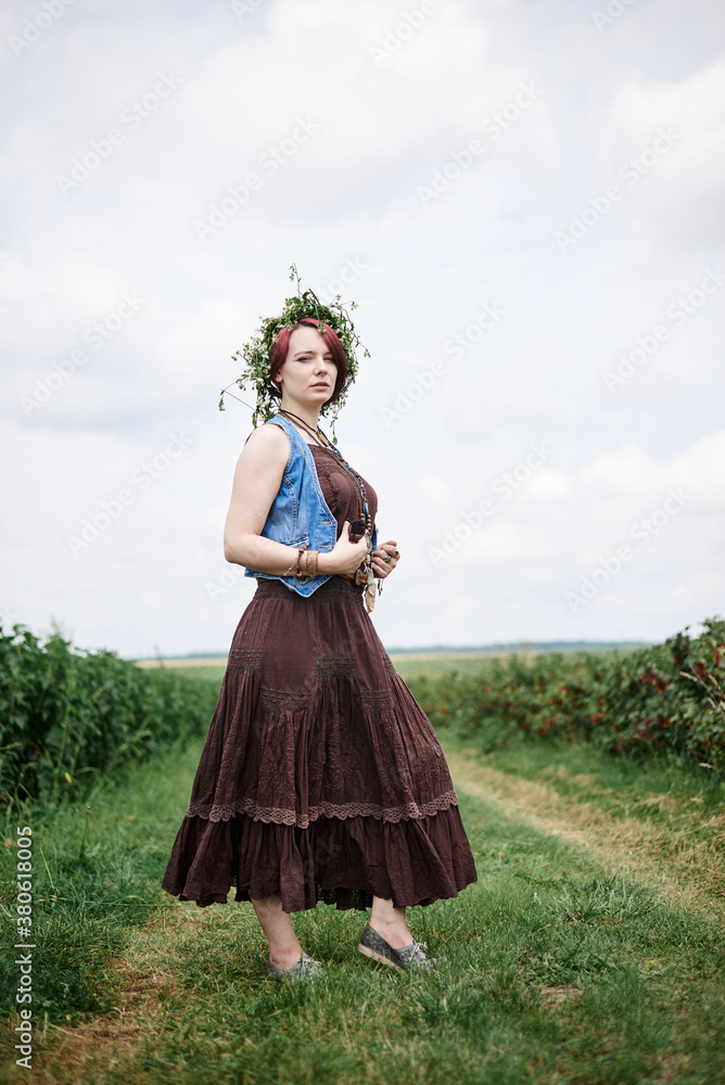Young hippie woman with short red hair, wearing boho style clothes and flower wreath, standing on green currant field, posing for picture. Female portrait on natural background. Eco tourism concept.