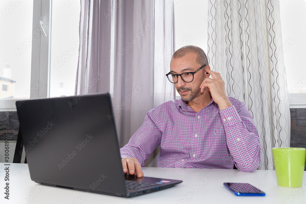 Adult bearded male wearing glasses and shirt sitting on a table using laptop to have a business meeting while drinks coffee with smartphone and pen. Working from home is the new normal concept. Adult bearded male wearing glasses and shirt sitting on a table using laptop to have a business meeting while drinks coffee with smartphone and pen. Working from home is the new normal concept.
