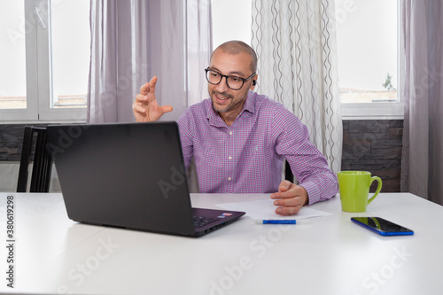 Adult bearded male wearing glasses and shirt sitting on a table using laptop to have a business meeting while drinks coffee with smartphone and pen. Working from home is the new normal concept.