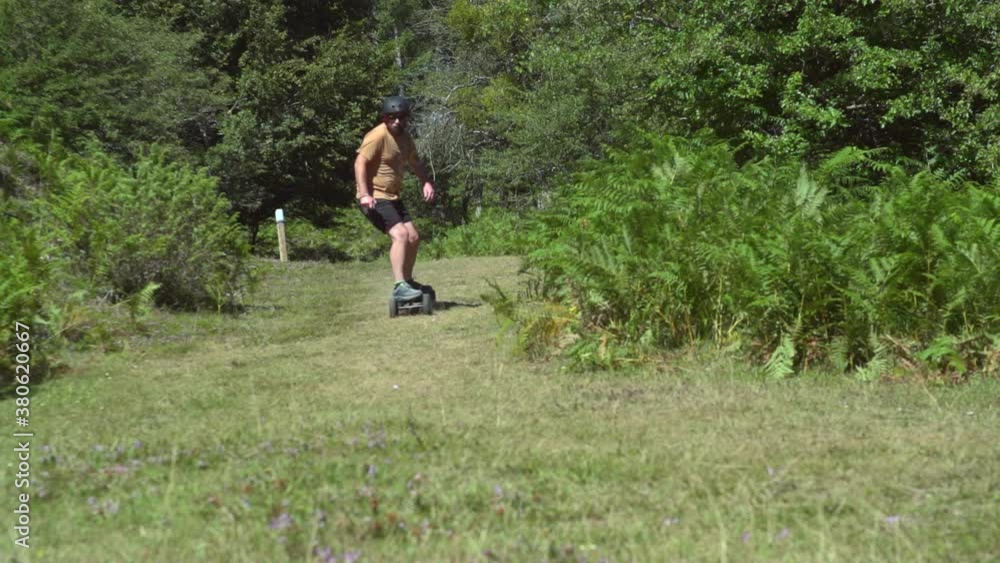 Adult Caucasian man in shorts rides self propelled remote control electric motorized longboard skateboard down outdoor sloping green grass hill in rural countryside landscape on sunny day, pan Adult Caucasian man in shorts rides self propelled remote control electric motorized longboard skateboard down outdoor sloping green grass hill in rural countryside landscape on sunny day, pan