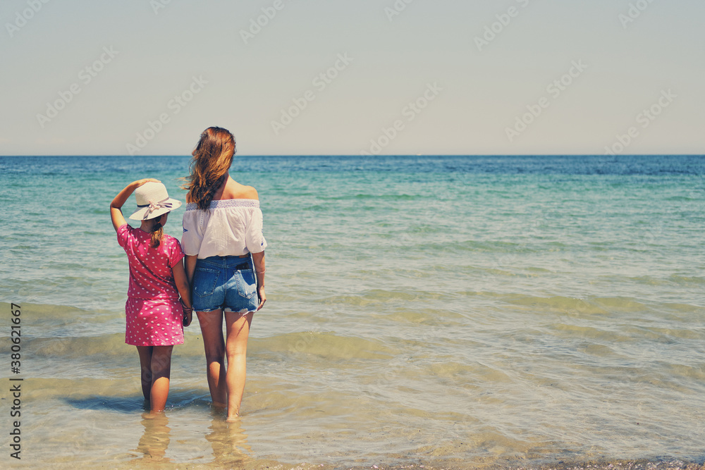 Back view mother and little daughter holding hands standing in sea water looking into horizon, copy space for text. Family travel and summer vacation concept Back view mother and little daughter holding hands standing in sea water looking into horizon, copy space for text. Family travel and summer vacation concept