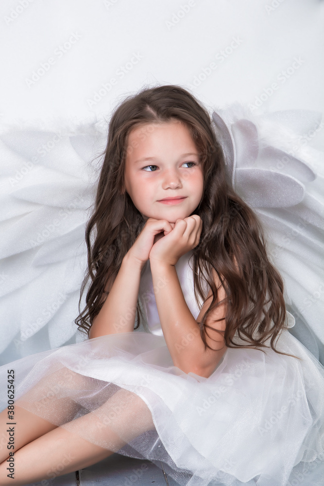 A cute dark-haired girl in a white dress with white angel wings. On a light background. Close-up. Place for an inscription.
A cute dark-haired girl in a white dress with white angel wings. On a light background. Close-up. Place for an inscription.