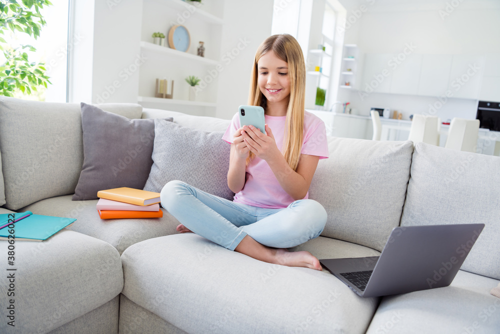Full length photo of positive kid girl study remote sit comfort couch legs crossed use smartphone comment social network post in house indoors Full length photo of positive kid girl study remote sit comfort couch legs crossed use smartphone comment social network post in house indoors