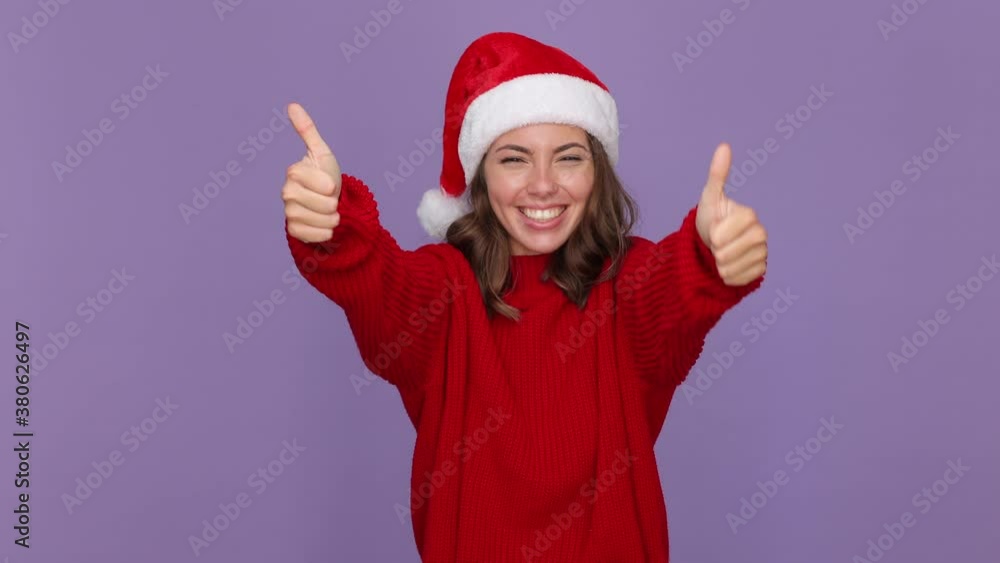 Cheerful young woman 20s in red knitted cozy sweater Santa Christmas hat showing thumbs up like gesture isolated on purple violet background studio. Happy New Year celebration merry holiday concept Cheerful young woman 20s in red knitted cozy sweater Santa Christmas hat showing thumbs up like gesture isolated on purple violet background studio. Happy New Year celebration merry holiday concept