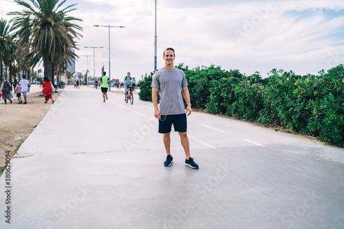 Portrait of cheerful muscular male runner standing on street enjoying workout and music in earphones, cheerful sportsman satisfied with online radio songs keeping healthy lifestyle and fit outdoors
