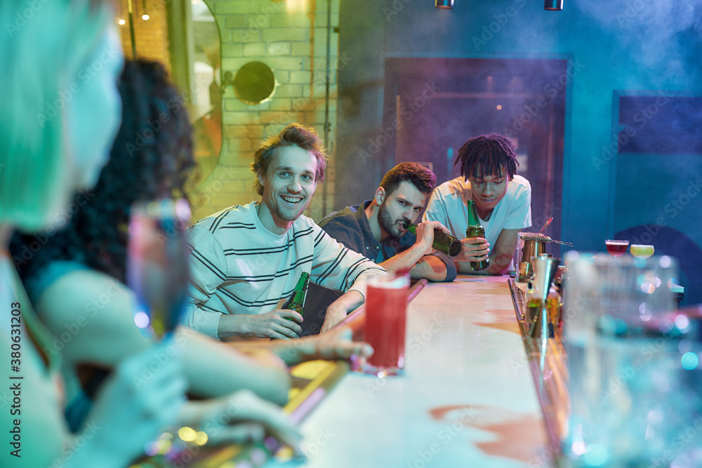 Make the night count. Three guys drinking beer, flirting, looking at women sitting at the bar counter. Friends spending time at night club, restaurant