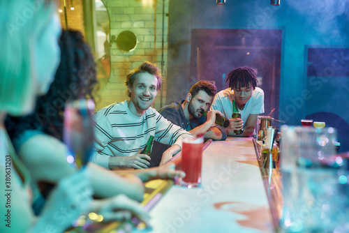 Make the night count. Three guys drinking beer, flirting, looking at women sitting at the bar counter. Friends spending time at night club, restaurant