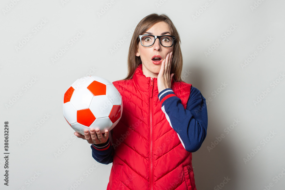 Young woman with a surprised face in a red vest holds a soccer ball in her hands on a light background Young woman with a surprised face in a red vest holds a soccer ball in her hands on a light background