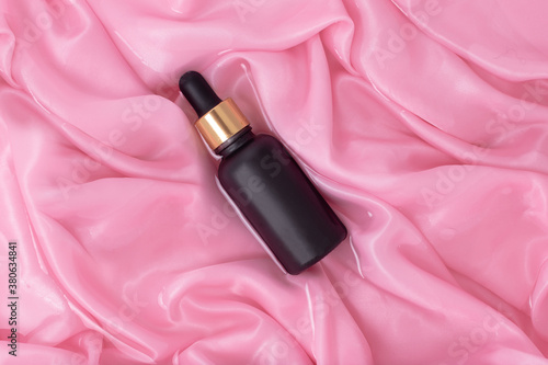 Matte black cosmetic bottle with dropper on a background of wet folds of pink silk. Mockup bottle for oil or essence.