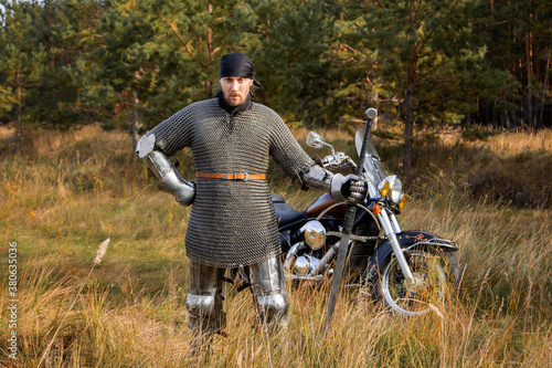 A medieval warrior in armor with a two-handed sword in his hands stands next to a motorcycle against the backdrop of a forest. Knight biker concept.