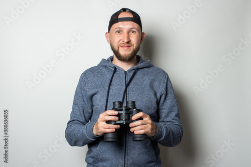 Young man in a hoodie and a cap holds binoculars in his hands on a light background