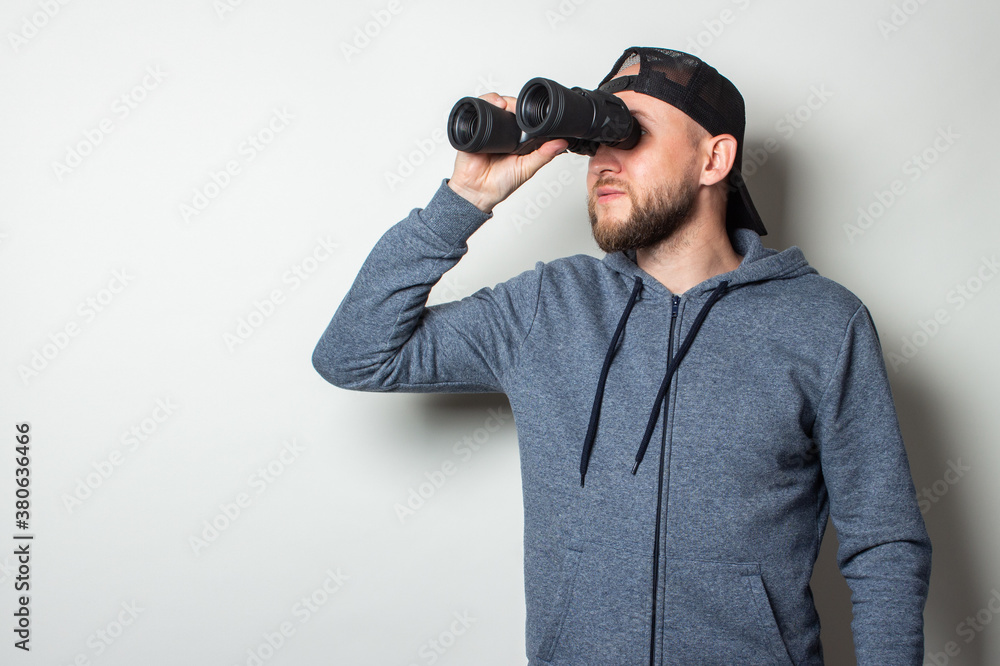 Young man in a hoodie and a cap looks through binoculars on a light background