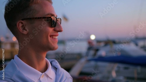 handsome man in white shirt walks along seafront near various motorboats against coloured sunset slow motion closeup