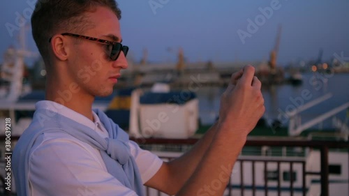 concentrated guy in black sunglasses makes photo of evening sunset seascape and smiles in summer slow motion closeup