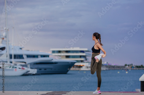Athlete and runner girl does stretching and warming up before or after a race.  It is right by the sea and you can see the boats docked in the port