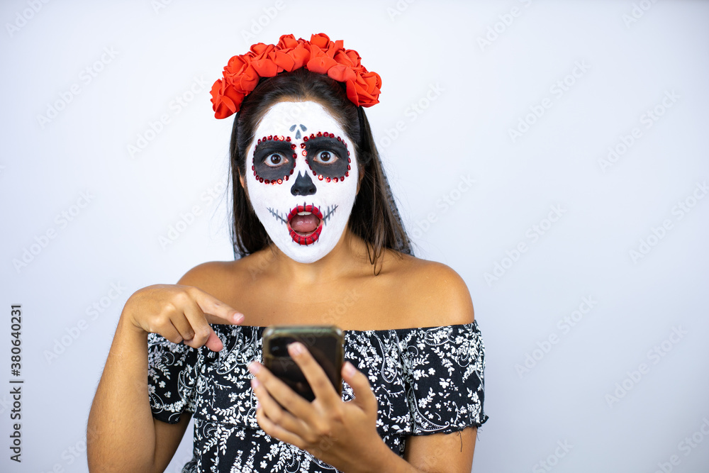 Woman wearing day of the dead costume over isolated white background chatting with her phone, surprised and pointing it