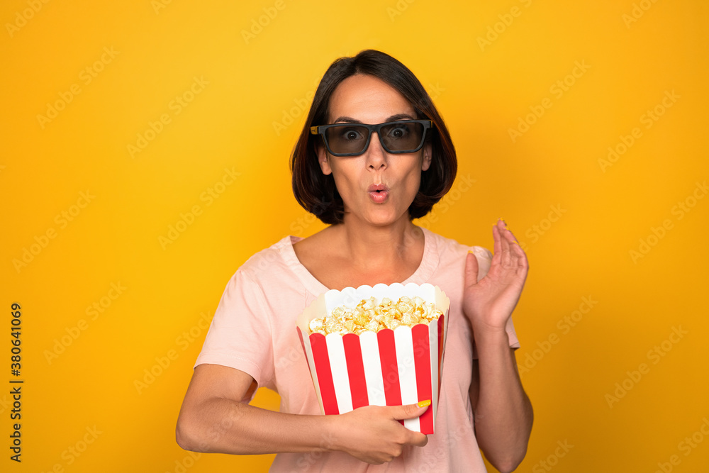 Woman in dark glasses holds box of popcorn. Young brunette with a surprised look raised her hand. Cut out on yellow background in studio. Cinema show concept. High quality photo. Woman in dark glasses holds box of popcorn. Young brunette with a surprised look raised her hand. Cut out on yellow background in studio. Cinema show concept. High quality photo.