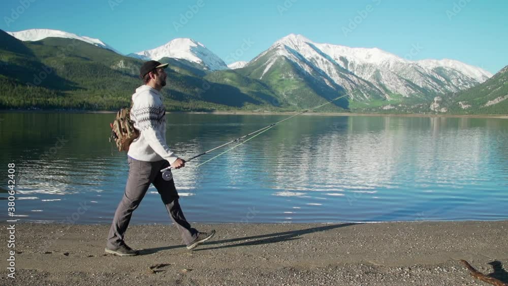 Wide tracking shot of a fly fisherman walking in front of gorgeous, snow-capped mountains, and a deep, reflective mountain lake. Shot at 240 fps. Wide tracking shot of a fly fisherman walking in front of gorgeous, snow-capped mountains, and a deep, reflective mountain lake. Shot at 240 fps.