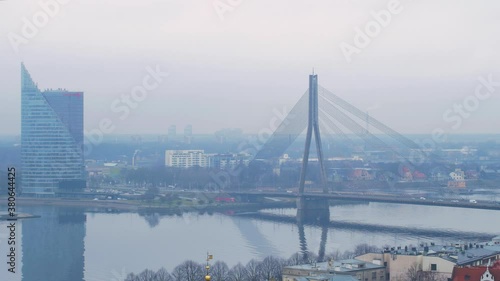 View on suspension bridge and modern skyscraper on the bank of Daugava river in Riga, overcast day with fog, wide high altitude rooftop cityscape shot photo