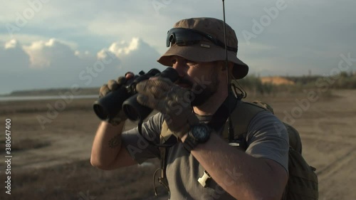 Close-up of guard patrolling plain area, soldier standing in military clothing, with backpack and radio transmitter, looking through binoculars and then report situation via walkie-talkie photo