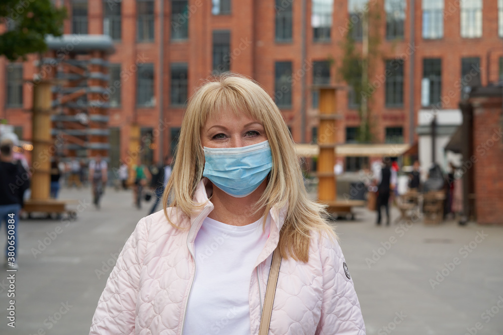 Woman in protective medical face mask standing outside in public place outdoor, looking at camera. Mature middle-aged female