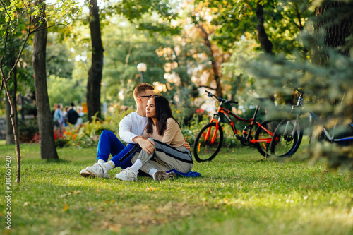 Beautiful young couple spends a weekend together in the park, sitting on the lawn and hugging on a bicycle background. Man and woman resting on the lawn in the autumn park after a bike ride.