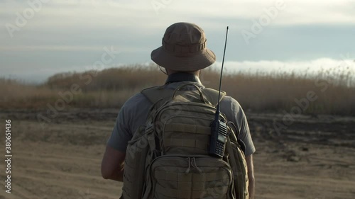 Slow motion rear view of bearded soldier, guard on duty with backpack and walkie-talkie, patrolling field, moving forward in plain area, checking territory for intruders photo