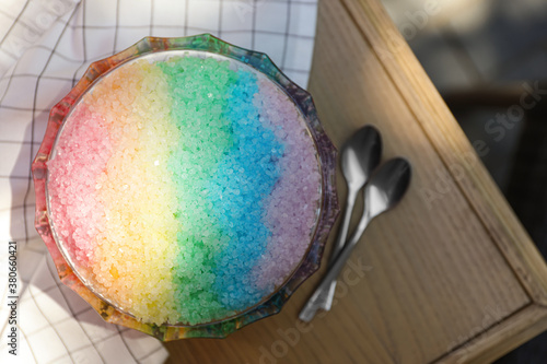 Rainbow shaving ice in glass dessert bowl and spoons on wooden table, flat lay