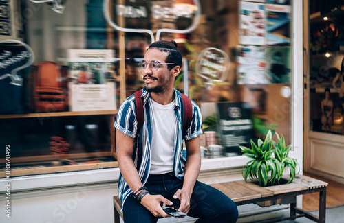 Cheerful dark skinned african american hipster guy in eyewear enjoying free time in city resting and using mobile phone, positive african american mali influencer holding smartphone and 4G #380670499