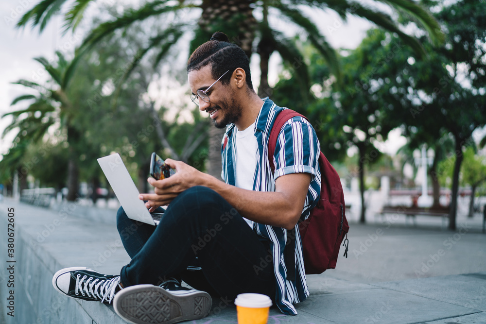 Happy young black hipster male using smartphone and laptop on city street